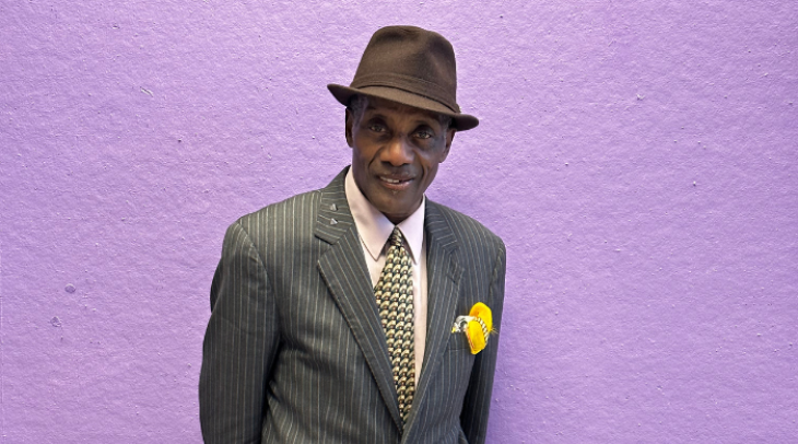 Elder black man in a gray suit, tie, and hat standing in front of a purple wall